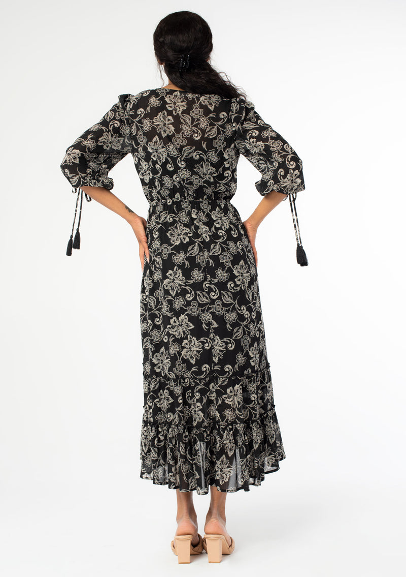 [Color: Black/Natural] A back facing image of a black model wearing a bohemian chiffon maxi dress in a black and natural floral print. With a high low ruffled hemline, a button front top, and three quarter length sleeves with tassel tie cuffs. 