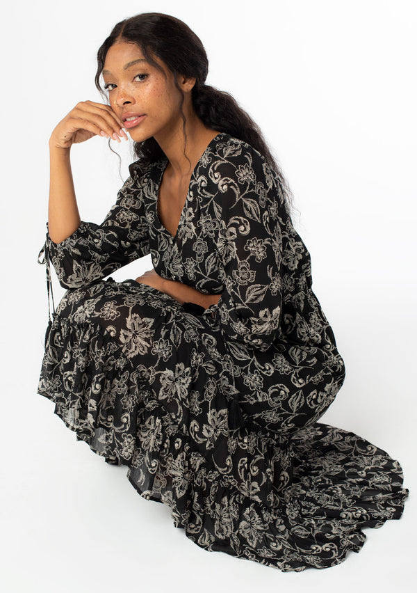 [Color: Black/Natural] A sitting image of a black model wearing a bohemian chiffon maxi dress in a black and natural floral print. With a high low ruffled hemline, a button front top, and three quarter length sleeves with tassel tie cuffs. 