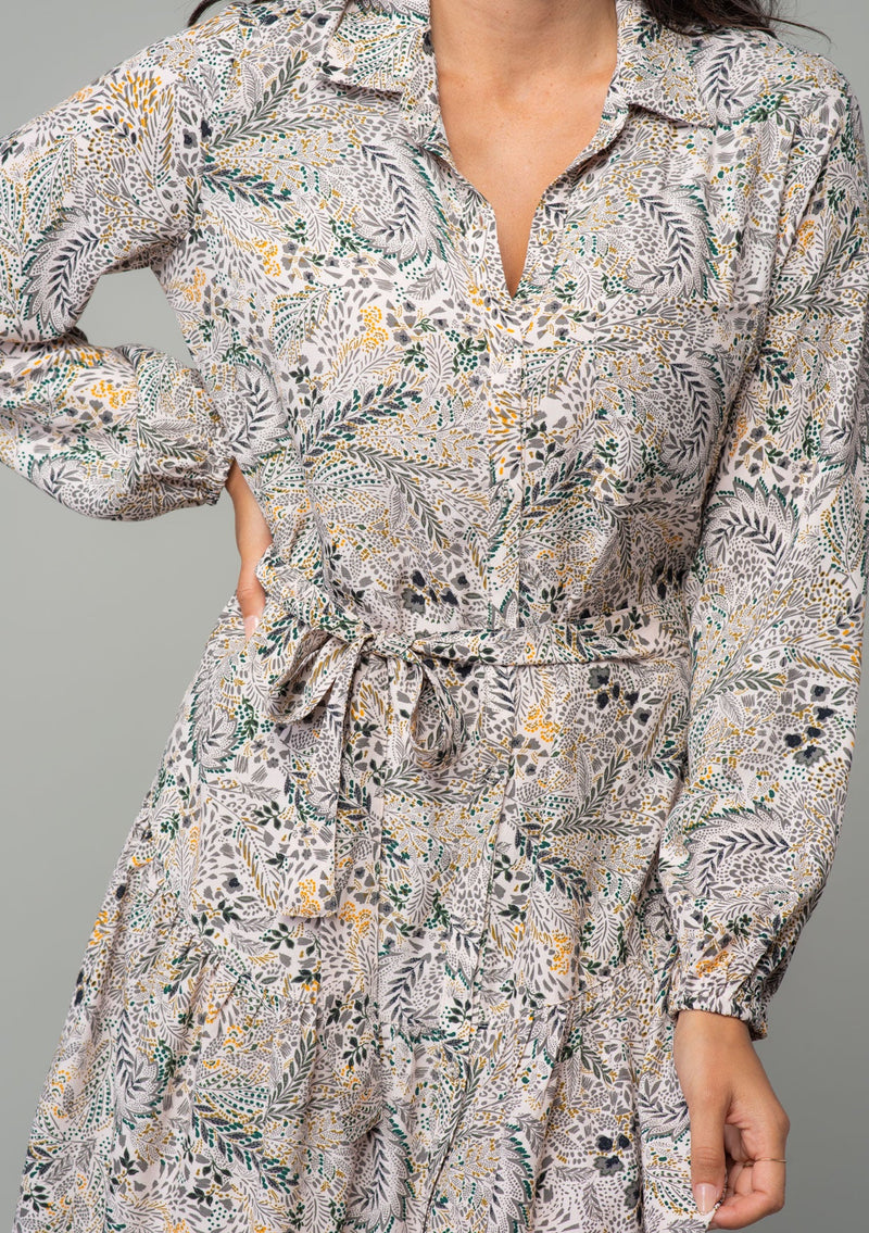 [Color: Natural/Olive] A close up front facing image of a brunette model wearing a natural and olive green floral print bohemian mini shirt dress. With long sleeves, a button front, a classic collared neckline, and a self tie waist belt. 