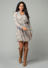 [Color: Natural/Olive] A front facing image of a brunette model wearing a natural and olive green floral print bohemian mini shirt dress. With long sleeves, a button front, a classic collared neckline, and a self tie waist belt. 