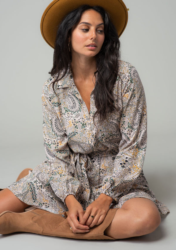 [Color: Natural/Olive] A front facing image of a brunette model wearing a natural and olive green floral print bohemian mini shirt dress. With long sleeves, a button front, a classic collared neckline, and a self tie waist belt. The model is sitting on the ground wearing a bohemian brown hat. 