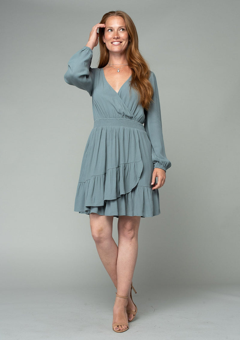 [Color: Balsam Green] A full body front facing image of a red headed model wearing a light green bohemian mini dress designed in a lightweight crepe. With long sleeves, a surplice v neckline, and a ruffled faux wrap skirt.