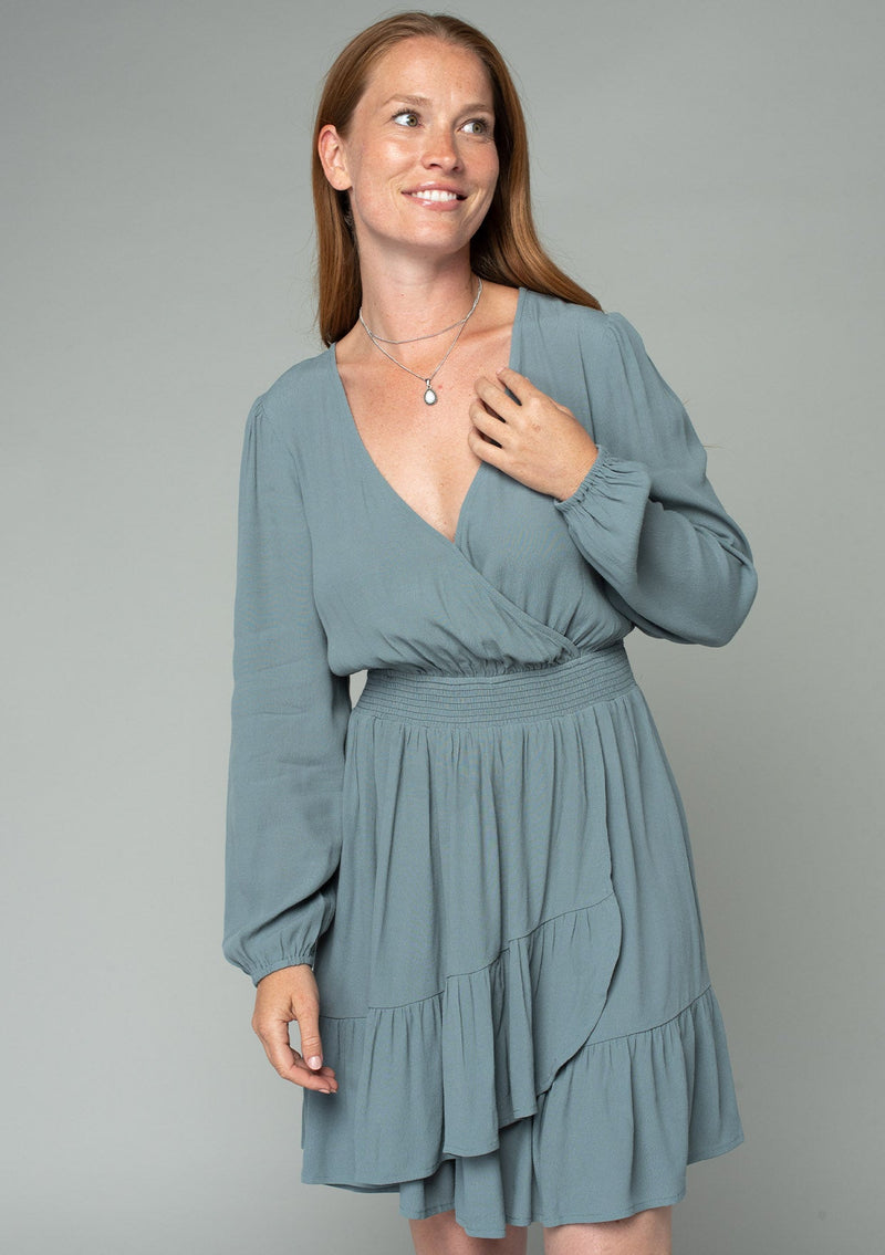 [Color: Balsam Green] A front facing image of a red headed model wearing a light green bohemian mini dress designed in a lightweight crepe. With long sleeves, a surplice v neckline, and a ruffled faux wrap skirt.