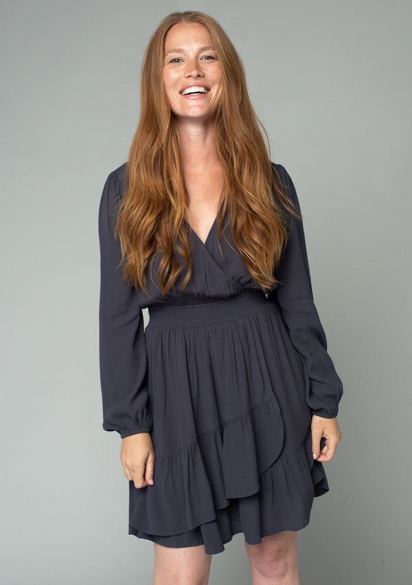 [Color: Charcoal] A front facing image of a red headed model wearing a charcoal grey bohemian mini dress designed in a lightweight crepe. With long sleeves, a surplice v neckline, and a ruffled faux wrap skirt.