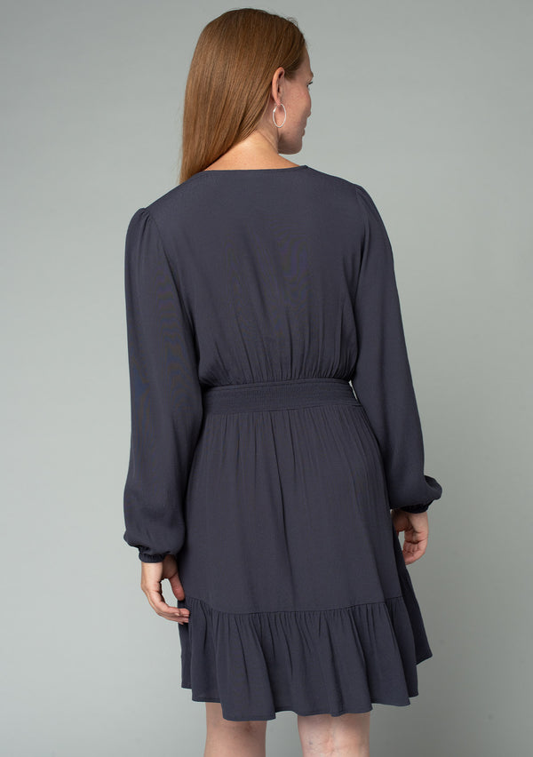 [Color: Charcoal] A back facing image of a red headed model wearing a charcoal grey bohemian mini dress designed in a lightweight crepe. With long sleeves, a surplice v neckline, and a ruffled faux wrap skirt. 