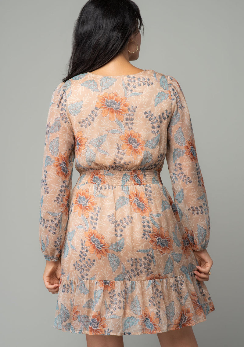 [Color: Coral/Blue] A back facing image of a brunette model wearing a sheer chiffon bohemian mini dress in a light coral and blue floral print. With long sleeves, a faux wrap front, and a ruffled hemline. 