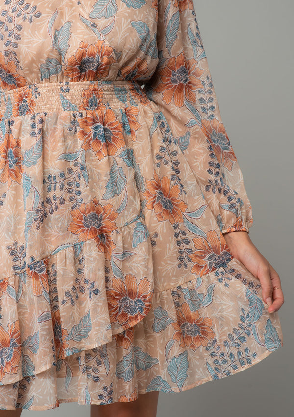 [Color: Coral/Blue] A close up front facing image of a brunette model wearing a sheer chiffon bohemian mini dress in a light coral and blue floral print. With long sleeves, a faux wrap front, and a ruffled hemline. 