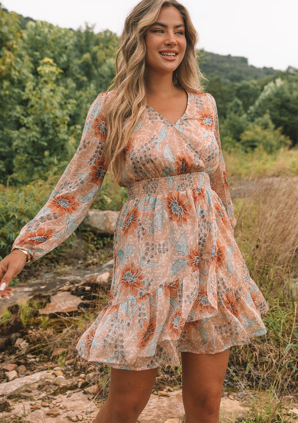 [Color: Coral/Blue] A front facing image of a blonde model wearing a sheer chiffon bohemian mini dress in a light coral and blue floral print. With long sleeves, a faux wrap front, and a ruffled hemline.