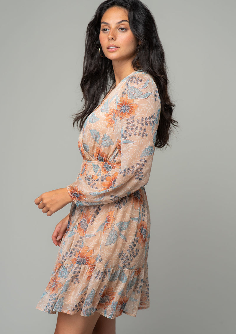 [Color: Coral/Blue] A side facing image of a brunette model wearing a sheer chiffon bohemian mini dress in a light coral and blue floral print. With long sleeves, a faux wrap front, and a ruffled hemline. 