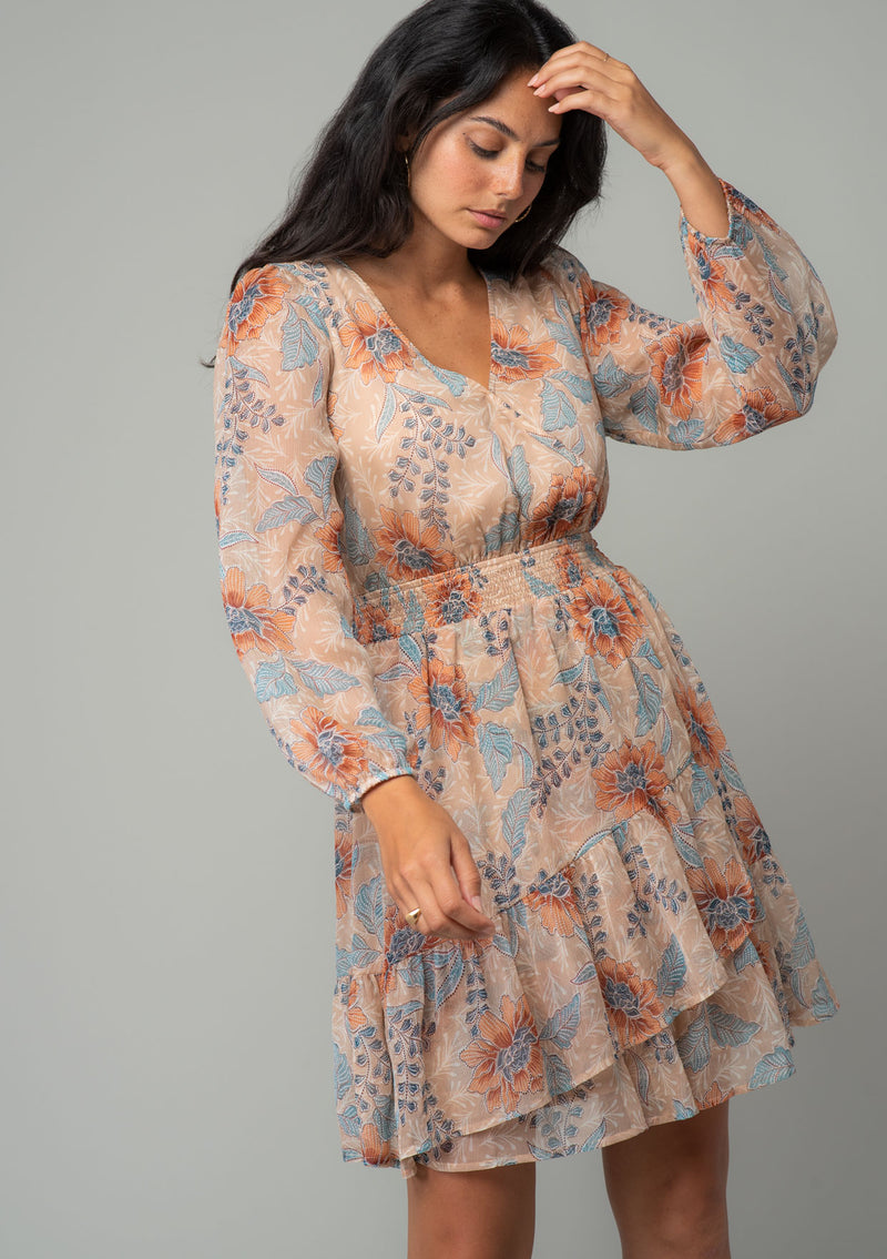 [Color: Coral/Blue] A front facing image of a brunette model wearing a sheer chiffon bohemian mini dress in a light coral and blue floral print. With long sleeves, a faux wrap front, and a ruffled hemline. The model has her arm up. 