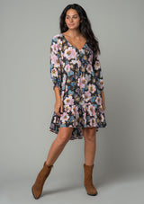 [Color: Charcoal/Rose] A full body front facing image of a brunette model wearing a charcoal grey and pink floral dress with a drawstring v neckline, a tiered high low hemline, and a flowy baby doll silhouette. 