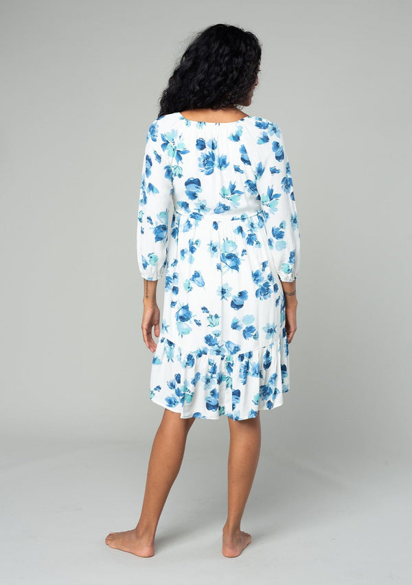 [Color: Ivory/Aqua] A back facing image of a brunette model wearing a flowy bohemian mini dress in an ivory white and blue floral print. With long sleeves, a tiered high low skirt, and a gathered v neckline with ties. 