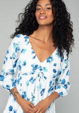 [Color: Ivory/Aqua] A close up front facing image of a brunette model wearing a flowy bohemian mini dress in an ivory white and blue floral print. With long sleeves, a tiered high low skirt, and a gathered v neckline with ties. 