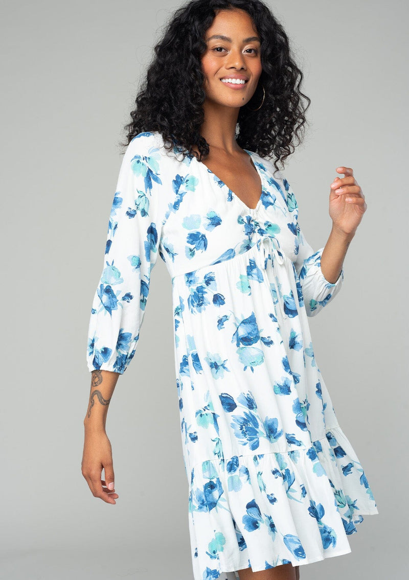 [Color: Ivory/Aqua] A side facing image of a brunette model wearing a flowy bohemian mini dress in an ivory white and blue floral print. With long sleeves, a tiered high low skirt, and a gathered v neckline with ties. 