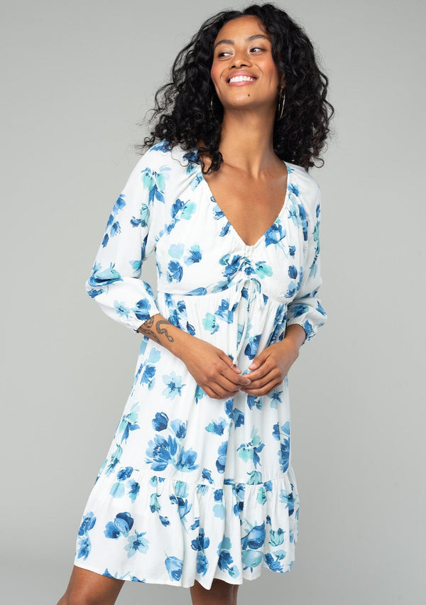[Color: Ivory/Aqua] A front facing image of a brunette model wearing a flowy bohemian mini dress in an ivory white and blue floral print. With long sleeves, a tiered high low skirt, and a gathered v neckline with ties. 