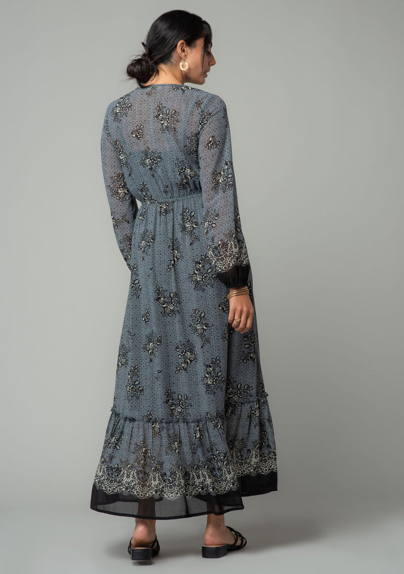 [Color: Blue/Black] A back facing image of a brunette model wearing a chiffon bohemian maxi dress in a blue and black floral border print. With long sleeves, a v neckline, and a tiered long skirt. 