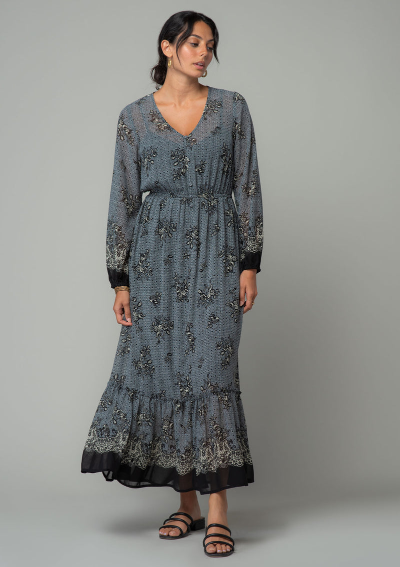 [Color: Blue/Black] A front facing image of a brunette model wearing a chiffon bohemian maxi dress in a blue and black floral border print. With long sleeves, a v neckline, and a tiered long skirt. 