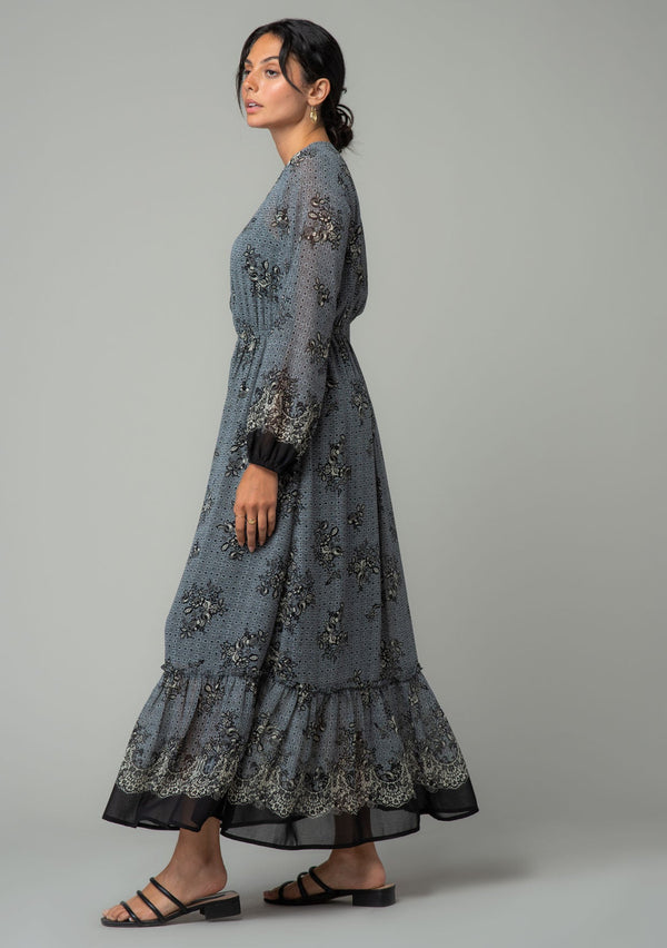 [Color: Blue/Black] A walking side facing image of a brunette model wearing a chiffon bohemian maxi dress in a blue and black floral border print. With long sleeves, a v neckline, and a tiered long skirt. 