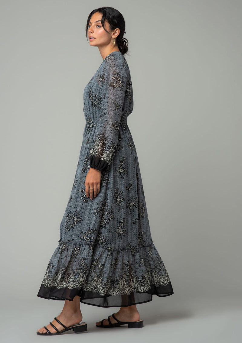 [Color: Blue/Black] A walking side facing image of a brunette model wearing a chiffon bohemian maxi dress in a blue and black floral border print. With long sleeves, a v neckline, and a tiered long skirt. 