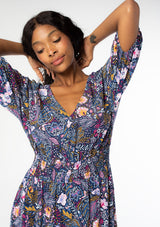 [Color: Navy/Berry] A close up front facing image of a black model wearing a navy blue and berry purple vintage floral print maxi dress with half length sleeves, a smocked elastic waist, and an open back with tassel ties. 