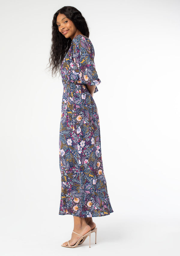 [Color: Navy/Berry] A side facing image of a black model wearing a navy blue and berry purple vintage floral print maxi dress with half length sleeves, a smocked elastic waist, and an open back with tassel ties. 