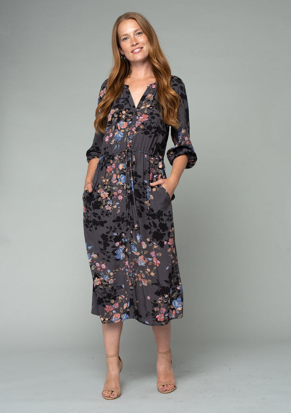 [Color: Grey/Dusty Blue] A front facing image of a red headed model wearing a lightweight bohemian mid length dress in a grey and blue floral print. With three quarter length sleeves, a button front, side pockets, and an adjustable drawstring waist. 