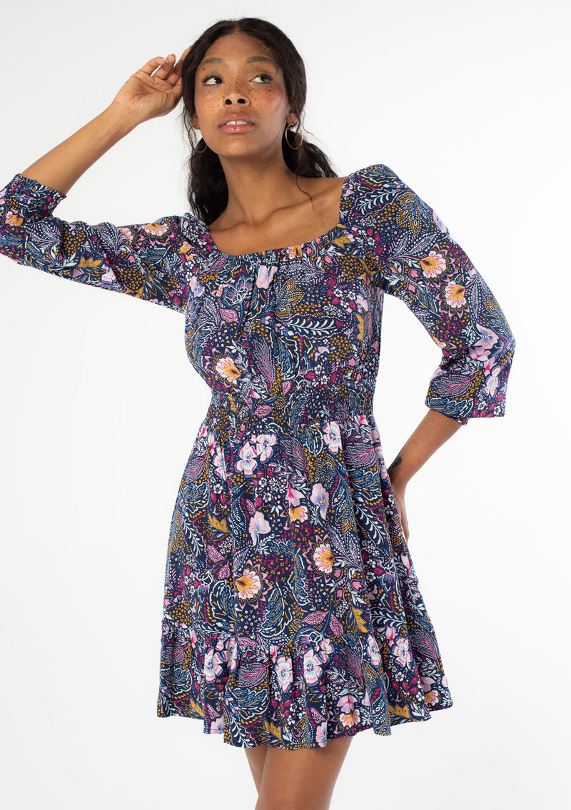 [Color: Navy/Berry] A front facing image of a black model wearing a navy blue and purple vintage floral print mini dress with long sleeves and a wide elastic neckline. 
