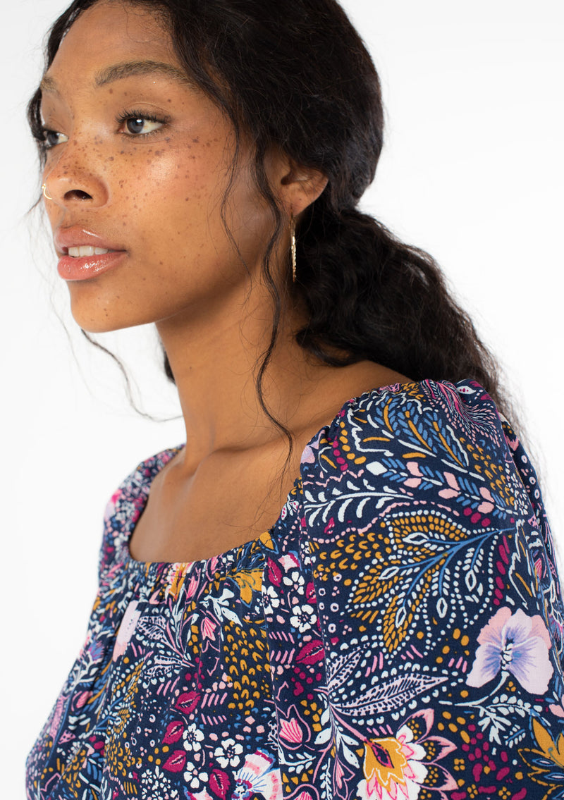 [Color: Navy/Berry] A close up side facing image of a black model wearing a navy blue and purple vintage floral print mini dress with long sleeves and a wide elastic neckline. 