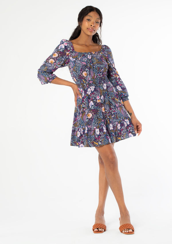 [Color: Navy/Berry] A full body front facing image of a black model wearing a navy blue and purple vintage floral print mini dress with long sleeves and a wide elastic neckline. 