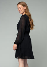 [Color: Black] A side facing image of a red headed model wearing a black mini dress in a clip dot chiffon. With sheer long sleeves, a flowy tiered skirt, an elastic waist, and a front waist tassel tie.
