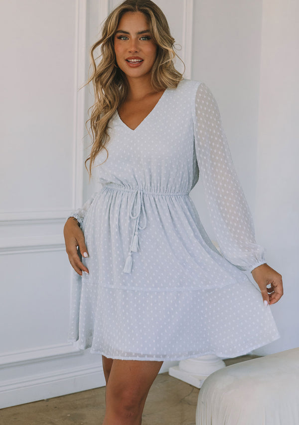[Color: Ice Blue] A front facing image of a blonde model wearing a light blue mini dress in a clip dot chiffon. With sheer long sleeves, a flowy tiered skirt, an elastic waist, and a front waist tassel tie.