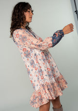 [Color: Ivory/Coral] A side facing image of a brunette model wearing a bohemian baby doll mini dress in an ivory white, coral pink, and navy blue mixed floral print. With voluminous long sleeves, a button front, and a tiered skirt. 
