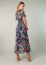 [Color: Navy/Rust] A back facing image of a blonde model wearing a sheer chiffon bohemian maxi dress in a navy blue and rust red floral print. Wit short flutter sleeves, a long tiered skirt, and an adjustable drawstring waist.