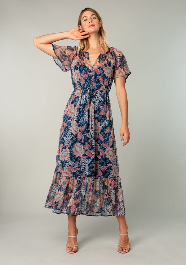 [Color: Navy/Rust] A front facing image of a blonde model wearing a sheer chiffon bohemian maxi dress in a navy blue and rust red floral print. Wit short flutter sleeves, a long tiered skirt, and an adjustable drawstring waist.