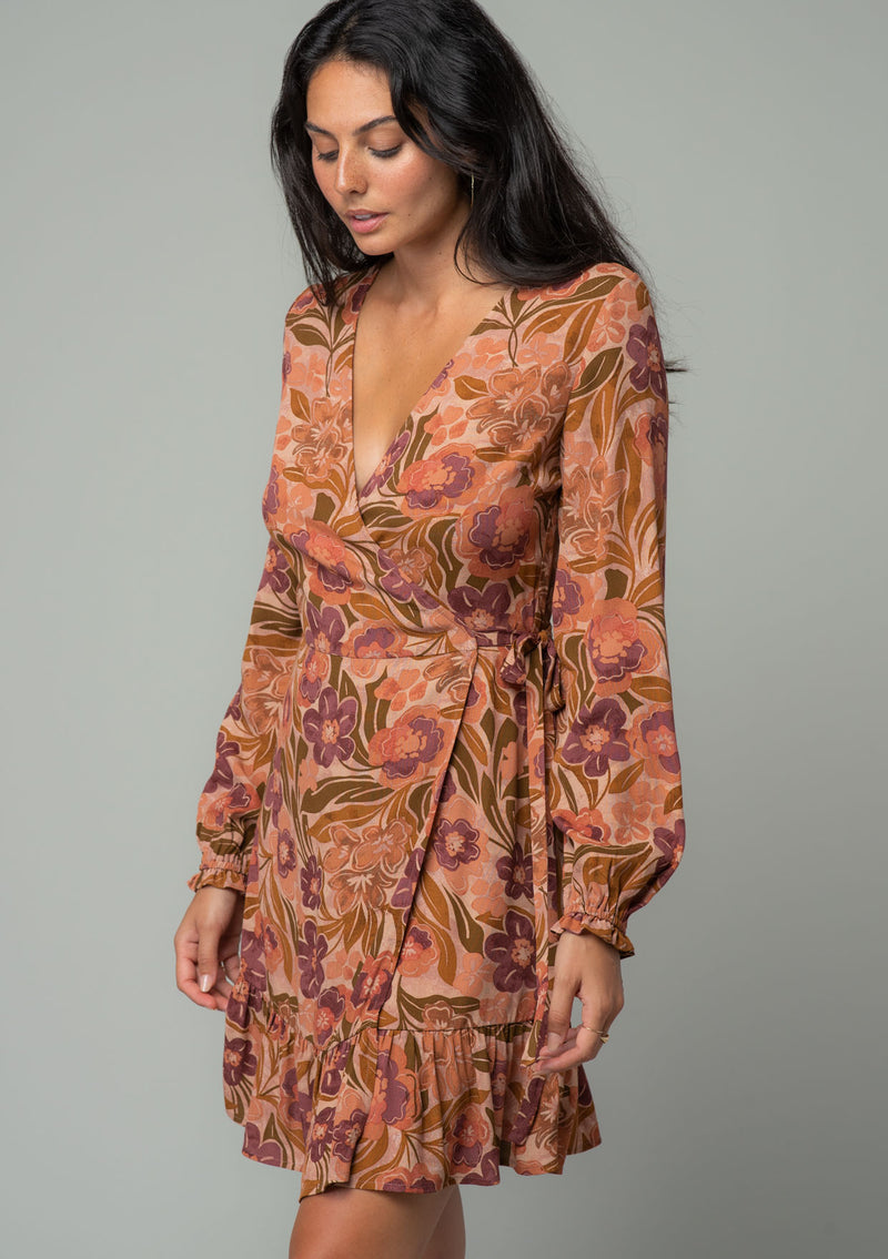 [Color: Clay/Olive] A side facing image of a brunette model wearing a clay brown and olive green retro floral print mini wrap dress. With long sleeves, a ruffled hemline, and a side tie closure. 