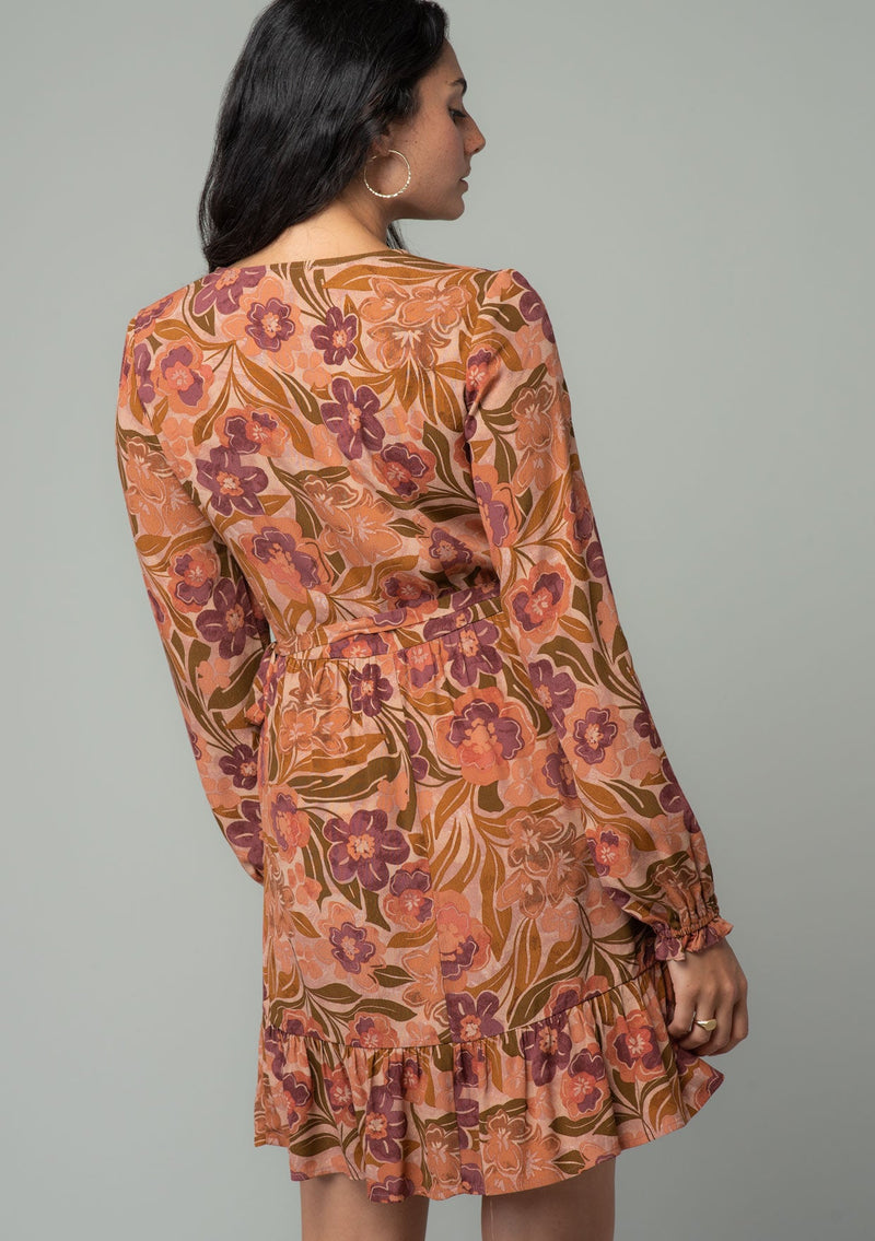 [Color: Clay/Olive] A back facing image of a brunette model wearing a clay brown and olive green retro floral print mini wrap dress. With long sleeves, a ruffled hemline, and a side tie closure. 