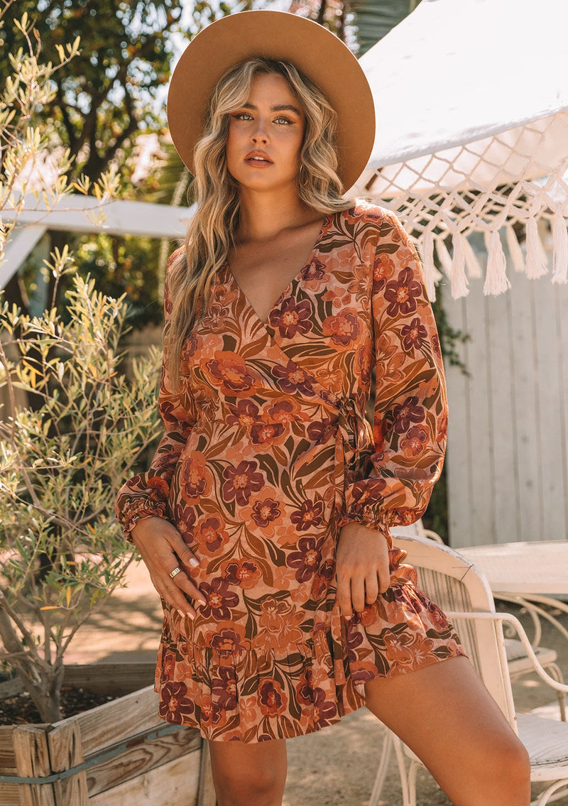 [Color: Clay/Olive] A front facing image of a blonde model wearing a clay brown and olive green retro floral print mini wrap dress. With long sleeves, a ruffled hemline, and a side tie closure.