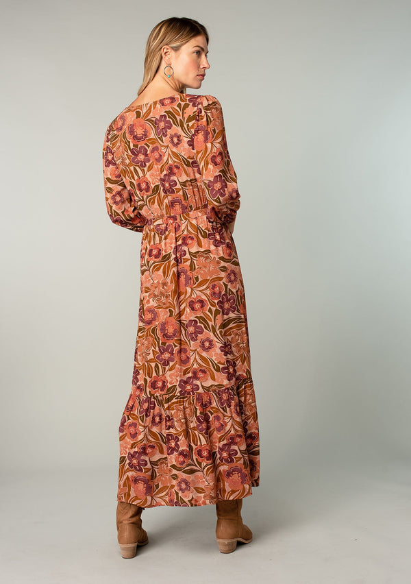 [Color: Clay/Olive] A back facing image of a blonde model wearing a clay brown and olive green retro floral print maxi dress. With long sleeves, a side slit, and a tie waist. 