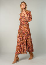 [Color: Clay/Olive] A front facing image of a blonde model wearing a clay brown and olive green retro floral print maxi dress. With long sleeves, a side slit, and a tie waist. 