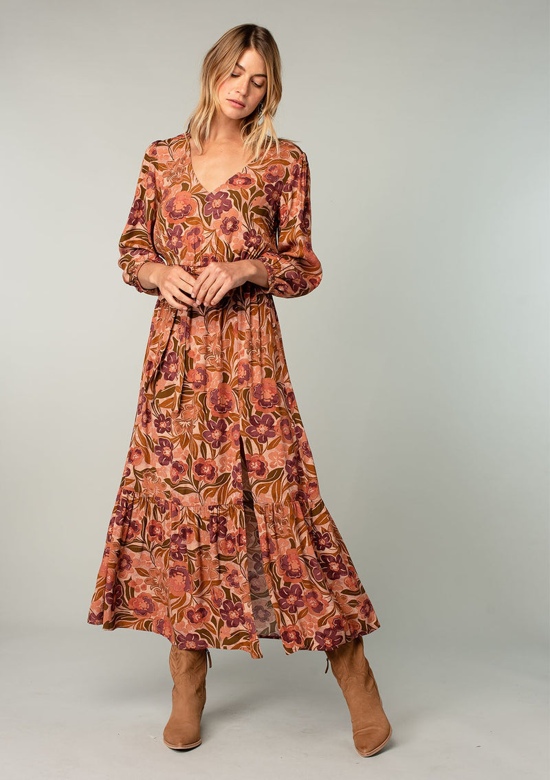 [Color: Clay/Olive] A front facing image of a blonde model wearing a clay brown and olive green retro floral print maxi dress. With long sleeves, a side slit, and a tie waist. 