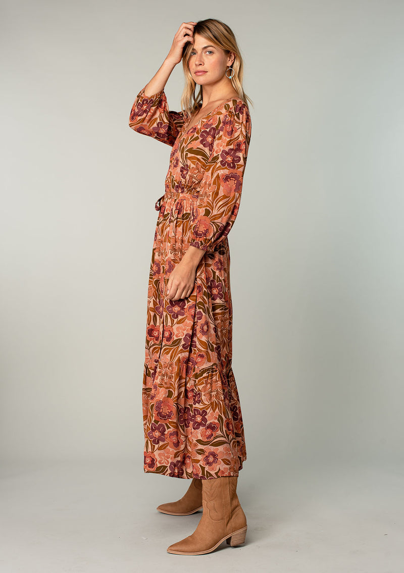 [Color: Clay/Olive] A side facing image of a blonde model wearing a clay brown and olive green retro floral print maxi dress. With long sleeves, a side slit, and a tie waist. 