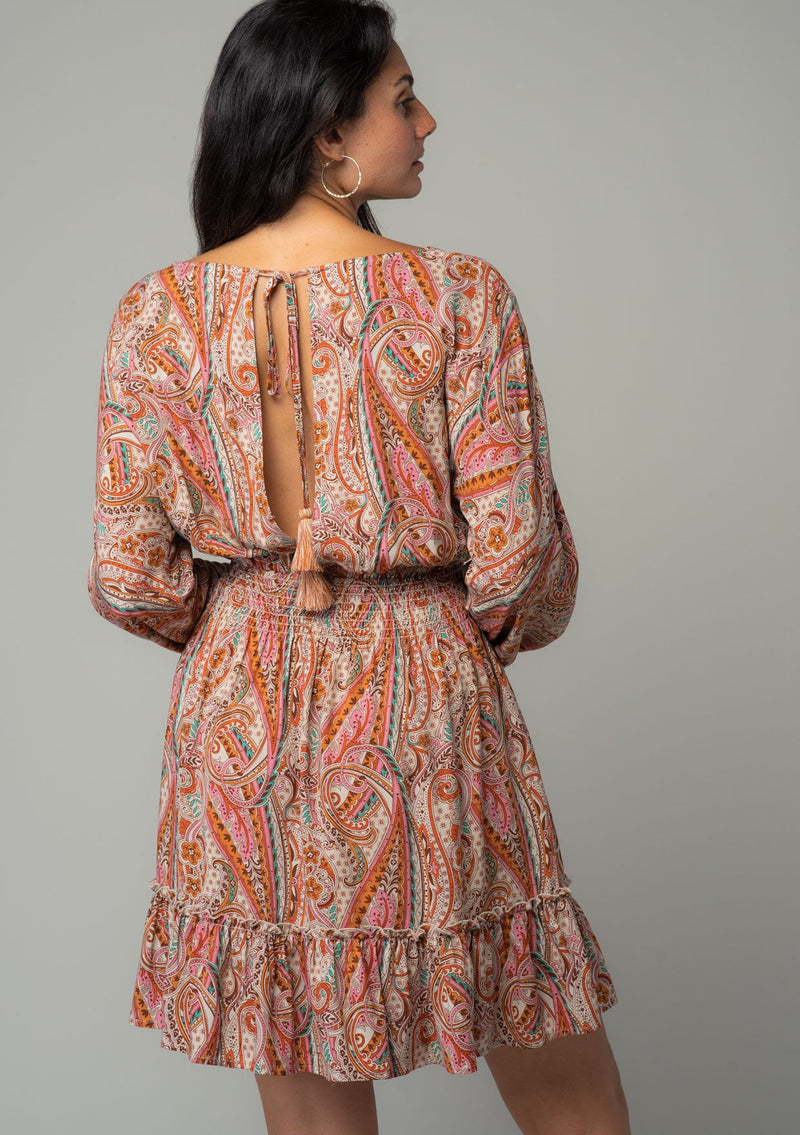 [Color: Natural/Rust] A back facing image of a brunette model wearing a bohemian mini dress in a natural and rust red paisley floral print. With long sleeves, a smocked elastic waist, and a back keyhole with tie closure. 