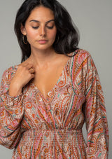[Color: Natural/Rust] A close up front facing image of a brunette model wearing a bohemian mini dress in a natural and rust red paisley floral print. With long sleeves, a smocked elastic waist, and a back keyhole with tie closure. 
