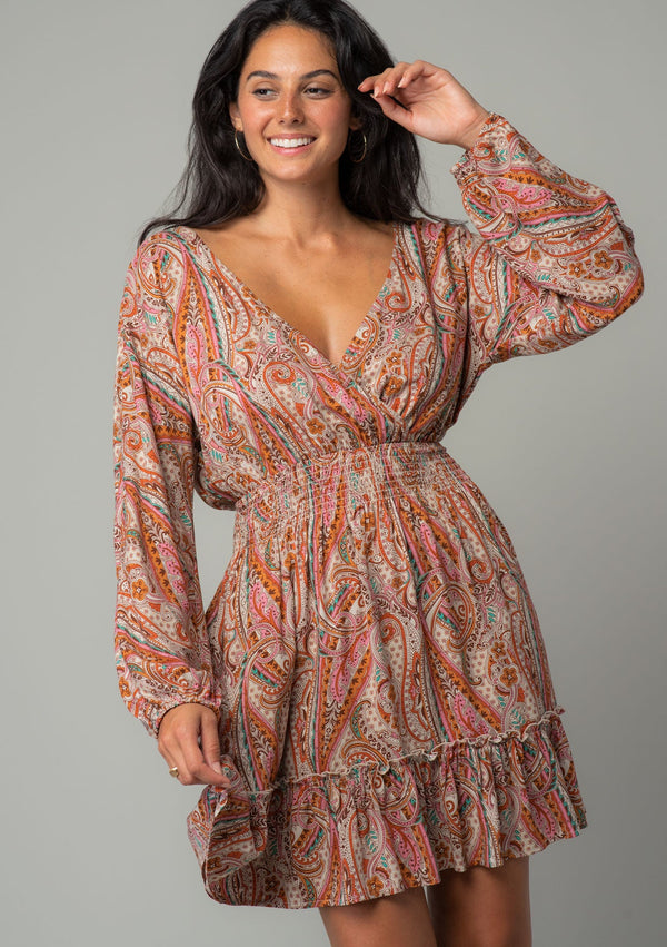 [Color: Natural/Rust] A front facing image of a brunette model wearing a bohemian mini dress in a natural and rust red paisley floral print. With long sleeves, a smocked elastic waist, and a back keyhole with tie closure. 