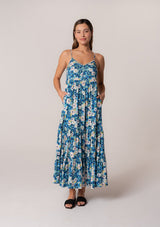 [Color: Dusty Teal/Blue] A front facing image of a brunette model wearing a flowy summer maxi dress in a blue floral print. With adjustable spaghetti straps, a scalloped edge v neckline, a button front top, a flowy tiered skirt, and side pockets. 