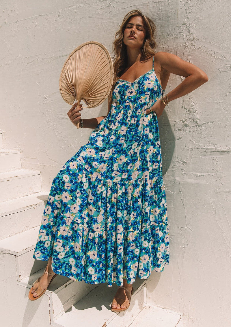 [Color: Dusty Teal/Blue] A front facing image of a blonde model standing outside holding a fan, wearing a flowy summer maxi dress in a blue floral print. With adjustable spaghetti straps, a scalloped edge v neckline, a button front top, a flowy tiered skirt, and side pockets. 