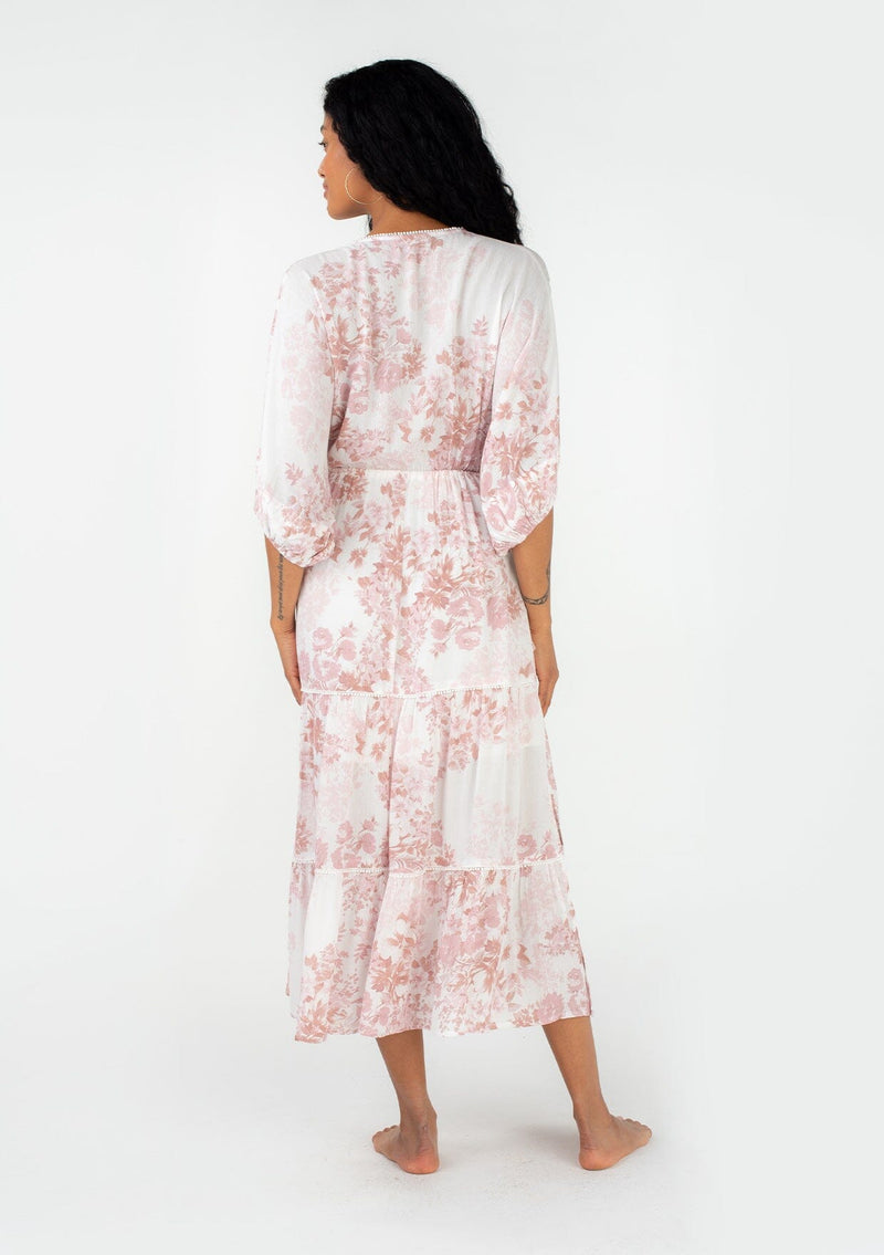 [Color: Ivory/Light Rust] A back facing image of a brunette model wearing a bohemian spring maxi dress in an ivory and light pink floral print. With half length puff sleeves, a surplice v neckline, a flowy tiered skirt, and a drawstring waistline with side tie details. Featuring little pom trim throughout. 