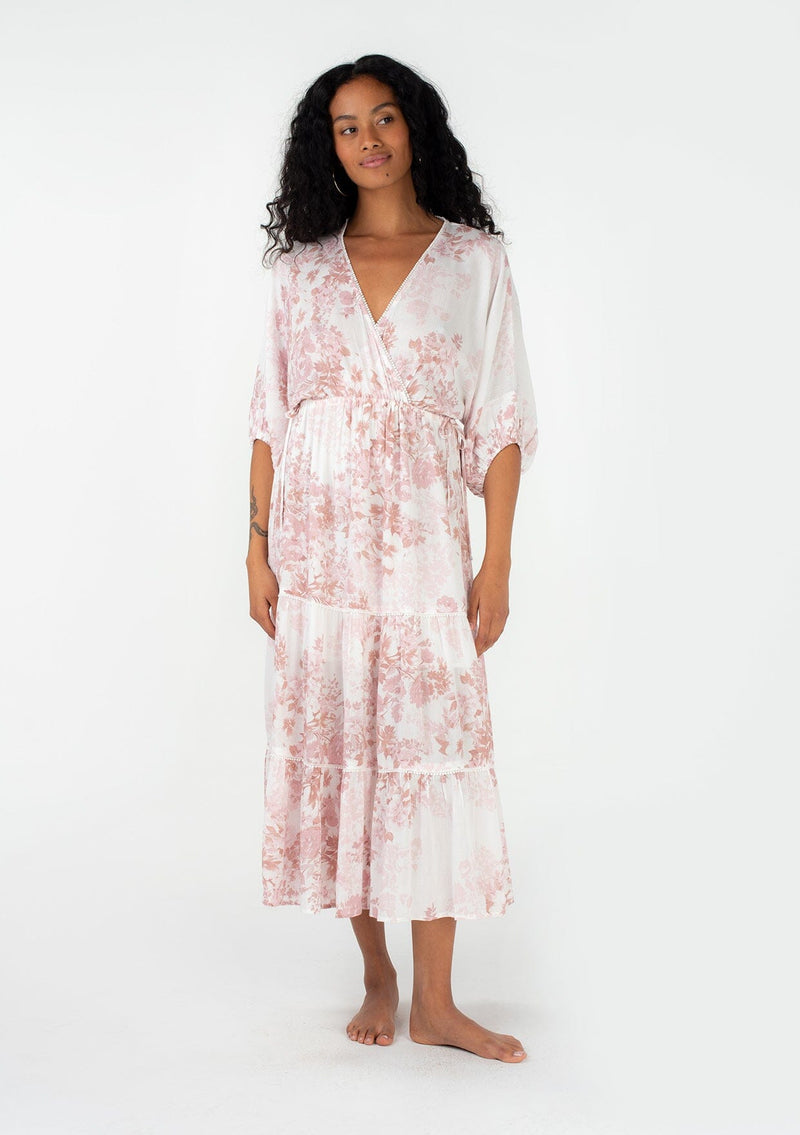 [Color: Ivory/Light Rust] A front facing image of a brunette model wearing a bohemian spring maxi dress in an ivory and light pink floral print. With half length puff sleeves, a surplice v neckline, a flowy tiered skirt, and a drawstring waistline with side tie details. Featuring little pom trim throughout. 