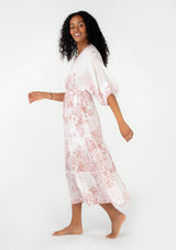 [Color: Ivory/Light Rust] A side facing image of a brunette model wearing a bohemian spring maxi dress in an ivory and light pink floral print. With half length puff sleeves, a surplice v neckline, a flowy tiered skirt, and a drawstring waistline with side tie details. Featuring little pom trim throughout. 