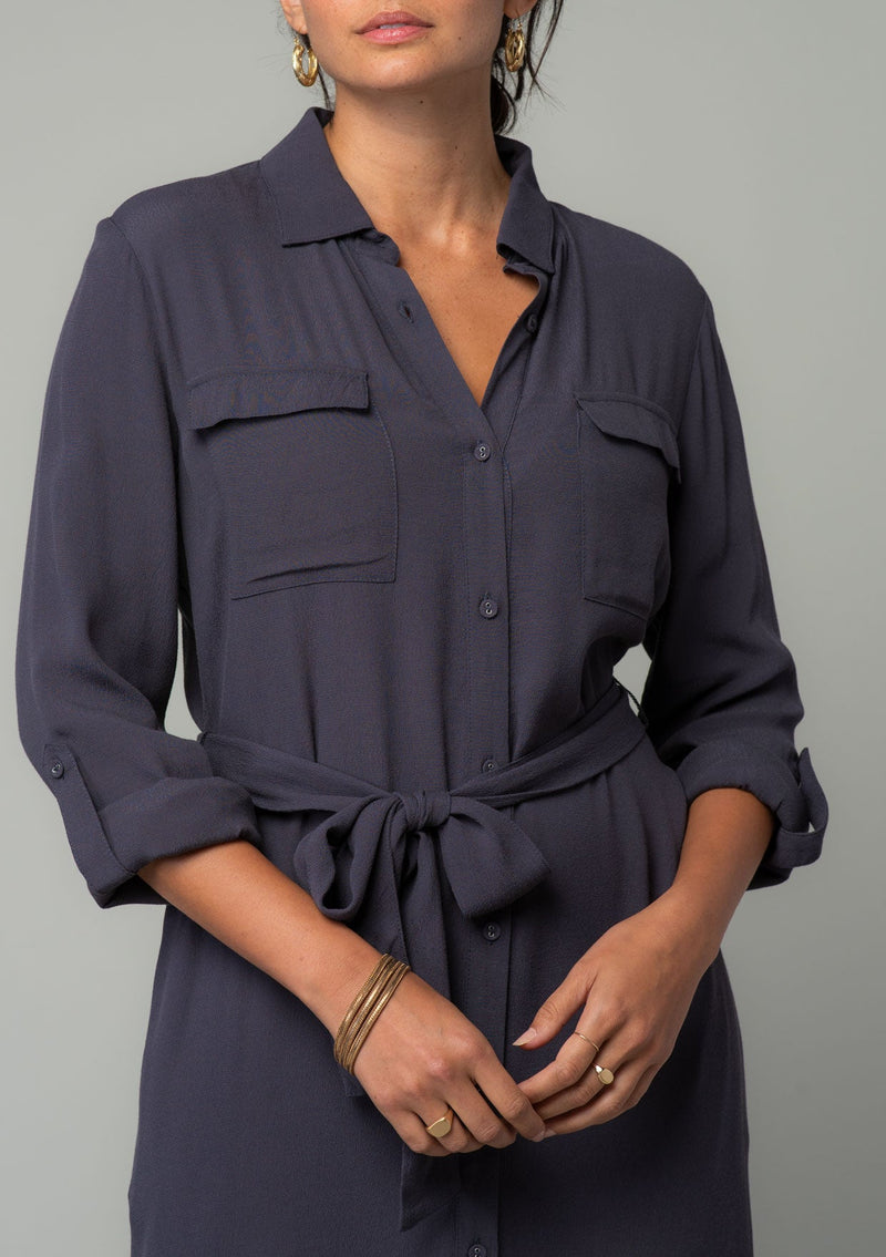 [Color: Charcoal] A close up front facing image of a brunette model wearing a relaxed fit charcoal grey maxi shirt dress made from a lightweight crepe. With long rolled sleeves, a button tab sleeve closure, two front pockets, side pockets at the hip, and a self tie waist belt. 